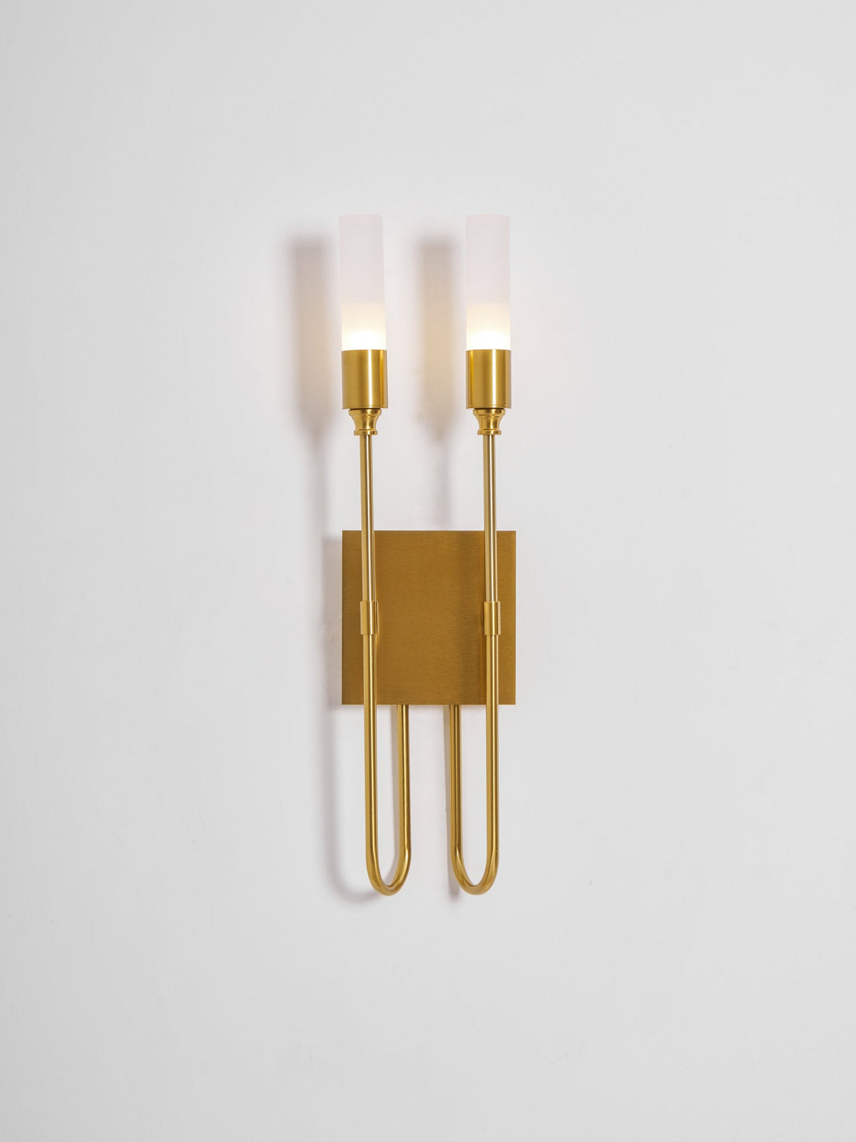 Brass two-light Clérachic sconce on a pale backdrop with both frosted tubes lit and the plate in warm light.