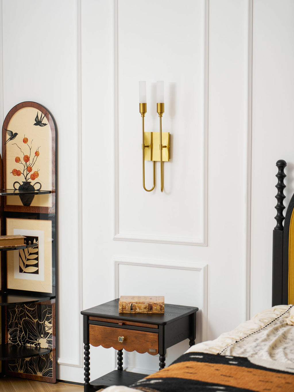 Small brass Clérachic two-light sconce on a white panelled wall, above a black nightstand and an arched painted cabinet.