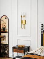 Small brass Clérachic two-light sconce on a white panelled wall, above a black nightstand and an arched painted cabinet.