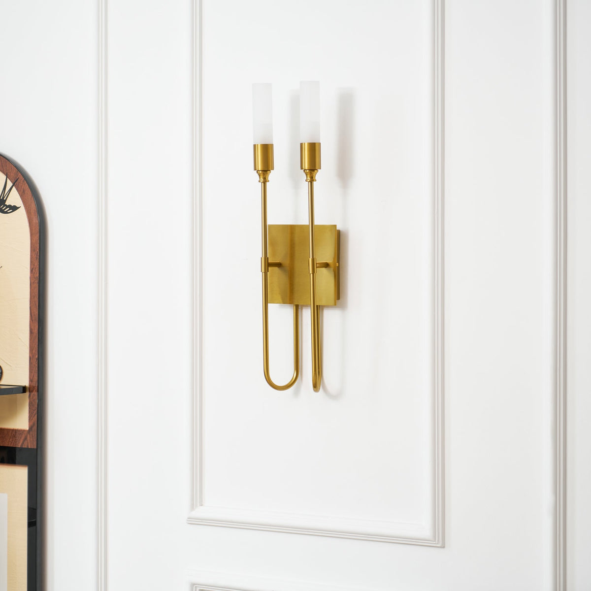 Close view of a brass two-light Clérachic sconce on a white panelled wall, the arched cabinet edge showing at the left.