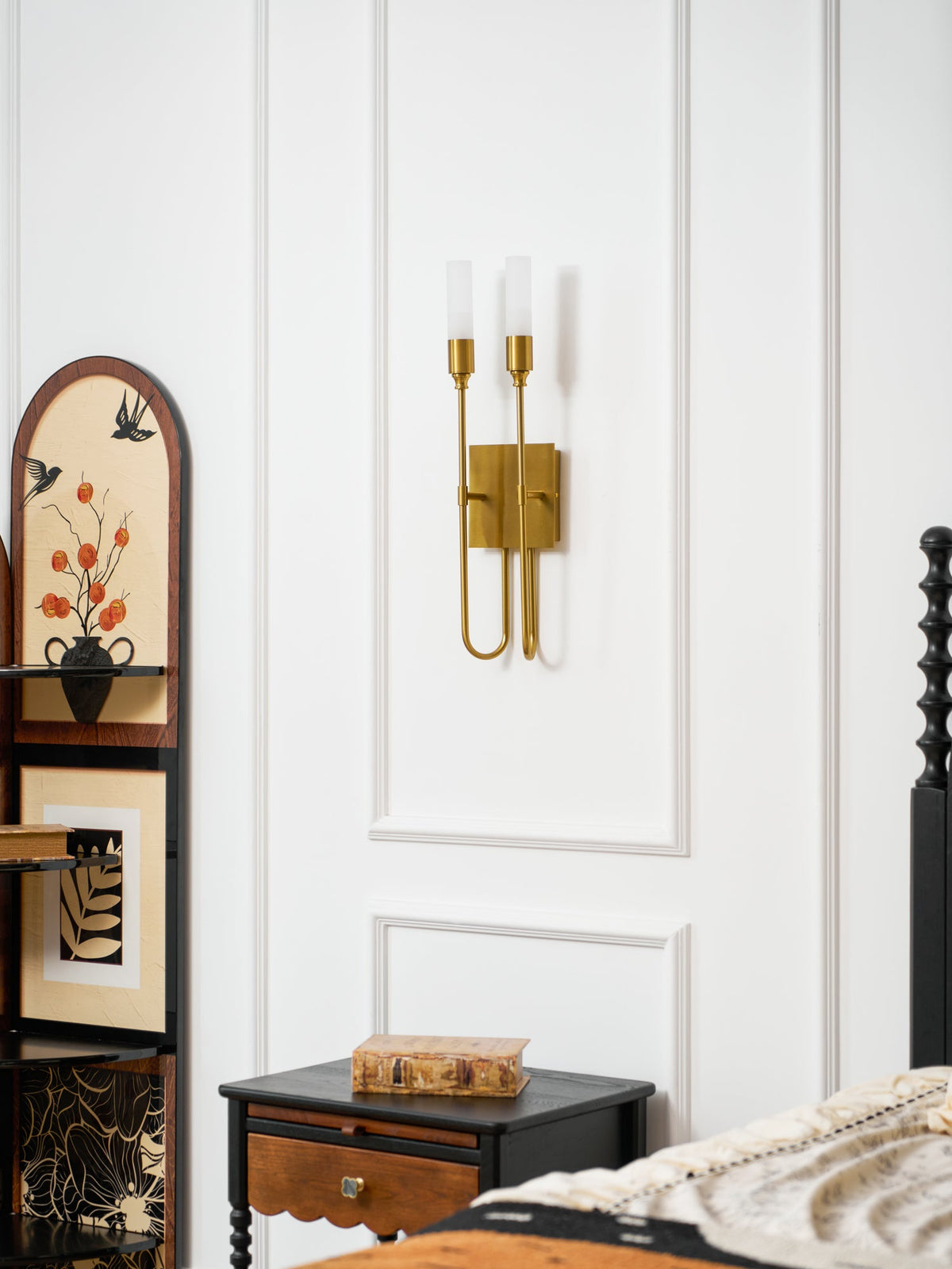 Brass two-light Clérachic sconce on white wall panelling, with a painted arched cabinet and a black nightstand below.