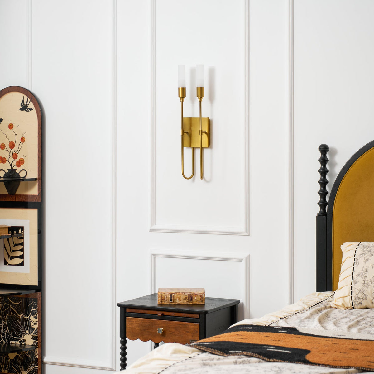 Brass Clérachic two-light sconce above a black nightstand with a brass-yellow velvet headboard at the right, on white panelling.