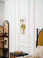 Angled view of a brass two-light Clérachic sconce on white panelling, with a bed and mustard headboard to the right.