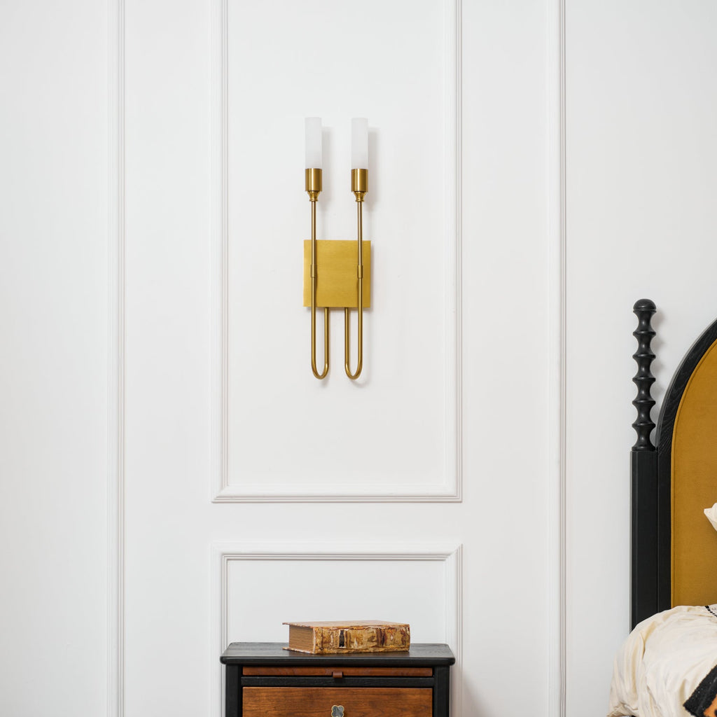 Straight-on brass two-light Clérachic sconce on a white panelled wall, above a nightstand and a black spindle bedpost.