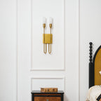Straight-on brass two-light Clérachic sconce on a white panelled wall, above a nightstand and a black spindle bedpost.
