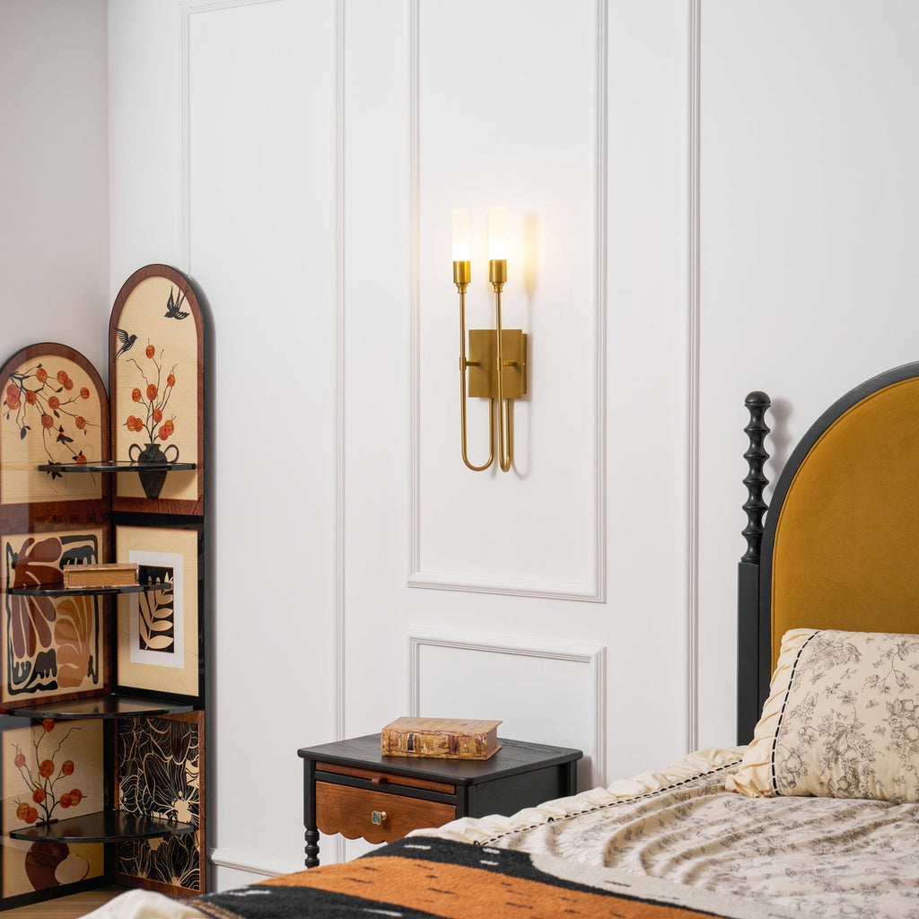 Lit brass two-light Clérachic sconce on a white panelled wall beside a bed with a mustard headboard and patterned bedding.
