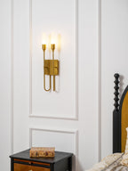 Brass two-light Clérachic sconce glowing on white wall panelling above a nightstand, with a black spindle bedpost at the right.