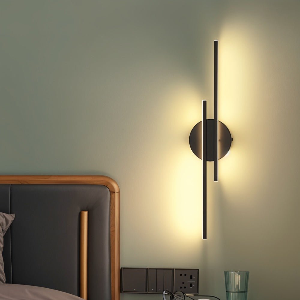 Clérachic black two-bar wall light glowing beside a bed with a wood-framed leather headboard, above a bedside socket panel