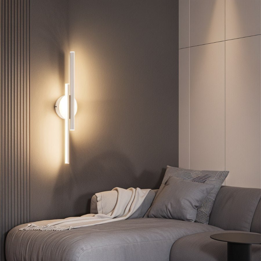 Clérachic white two-bar wall light glowing softly above a grey sofa with a throw, against a ribbed taupe wall