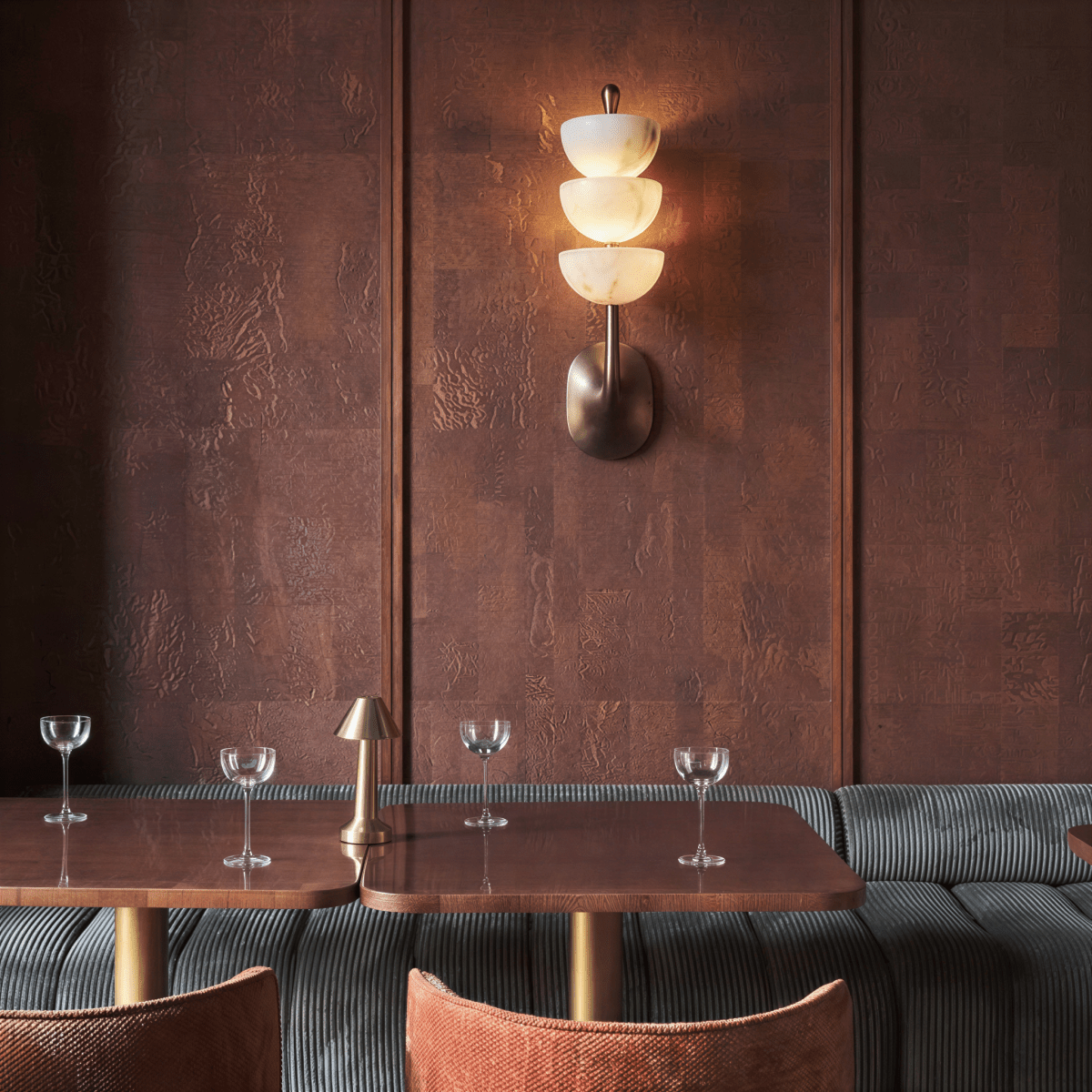 Clérachic tiered wall sconce glowing on a textured rust-toned panelled wall above a restaurant booth, with stemware and a small table lamp on the wooden tables below.