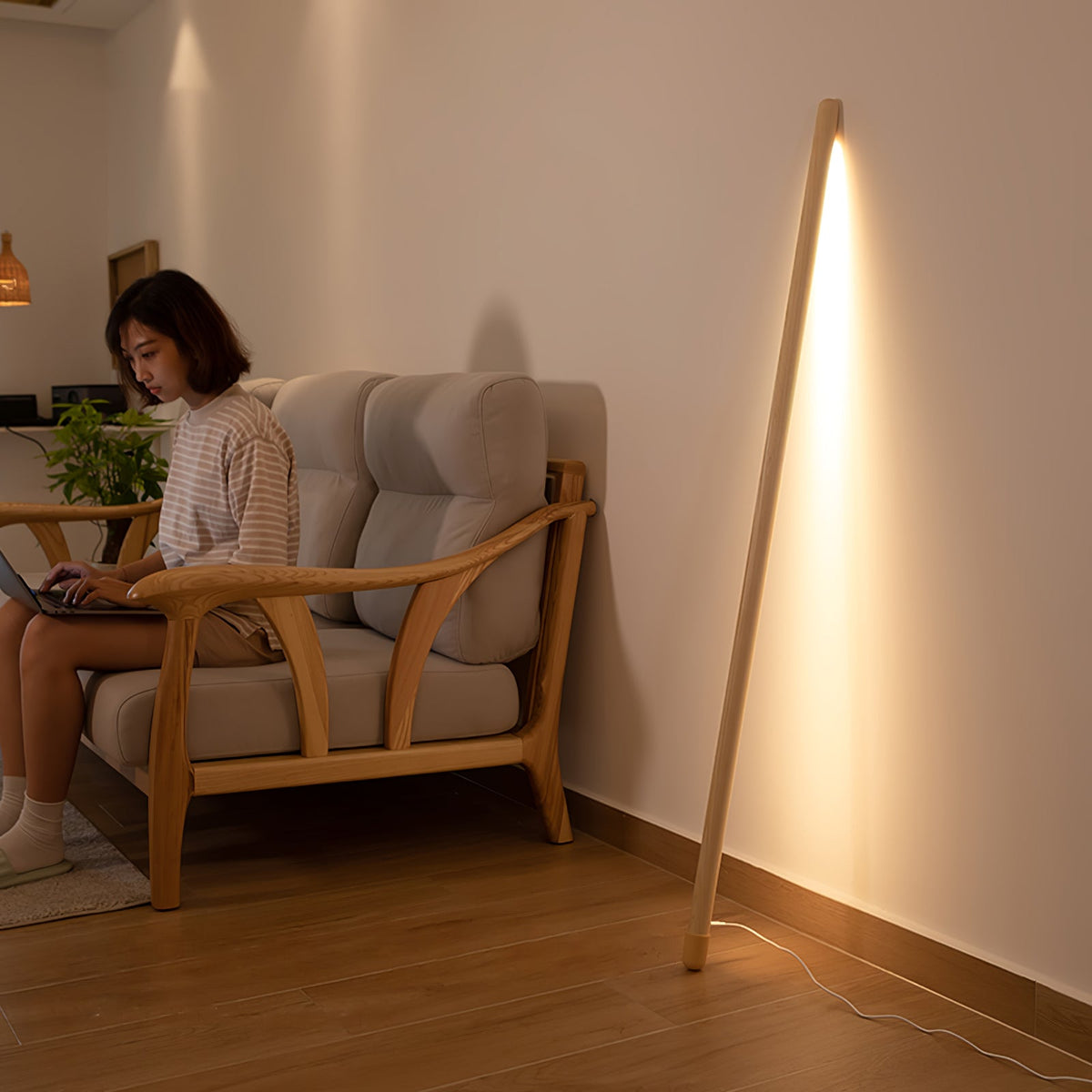 Light wood Clérachic floor lamp leaning in a corner and glowing warm, beside a woman working on a laptop in an armchair.
