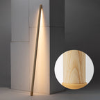 A light wood Clérachic floor lamp leaning against a grey wall corner, lit warm, with a round inset of its pale wood grain and base.