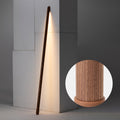Walnut-colour Clérachic floor lamp leaning in a corner and lit warm, with a round inset showing the grain and rounded foot.