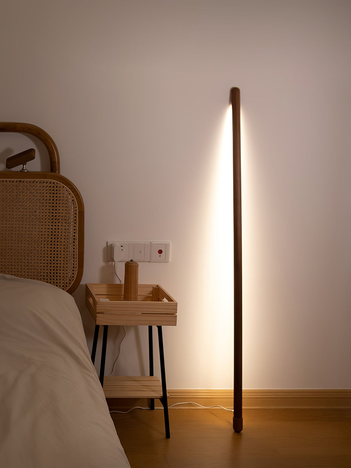Lit walnut-colour Clérachic floor lamp standing by a bed with a rattan headboard and a slim wooden bedside table.