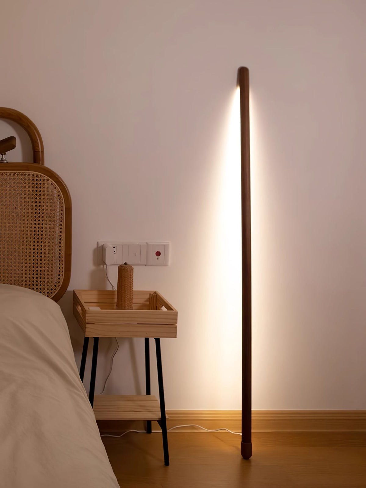 Walnut-coloured Clérachic floor lamp glowing beside a bed, with a rattan headboard and a small open shelf table.