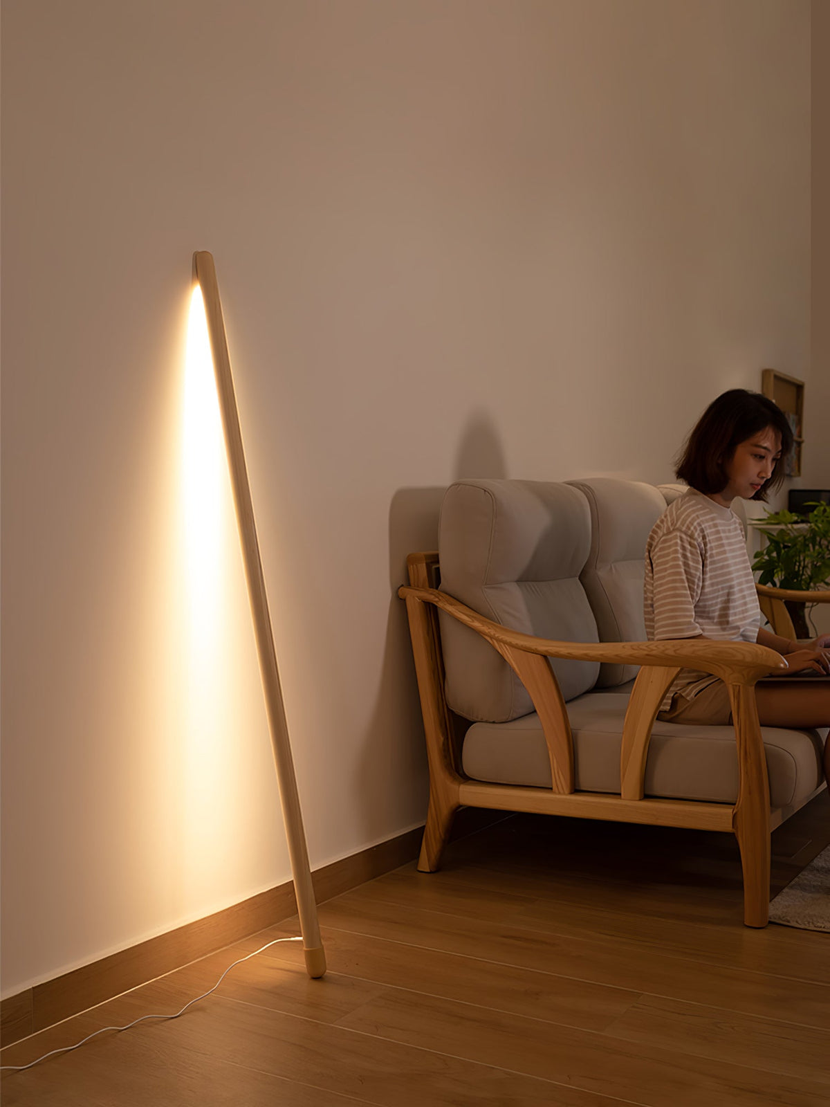 Light wood Clérachic floor lamp leaning against a beige wall and glowing warm, with a person seated in a wooden armchair.