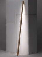 Light wood Clérachic floor lamp leaning in a grey wall corner, lit in a bright white and casting a long strip of light.