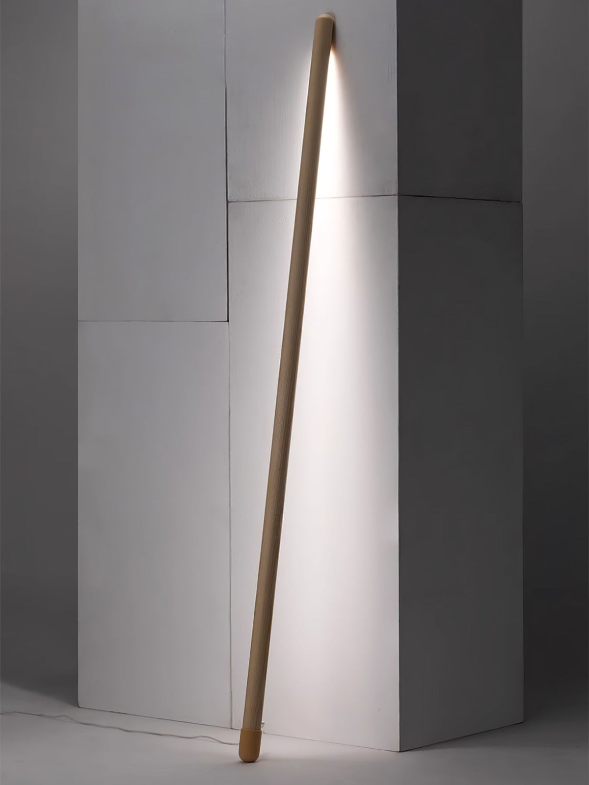 Light wood Clérachic floor lamp leaning in a grey wall corner, lit in a bright white and casting a long strip of light.