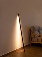 Walnut-colour Clérachic floor lamp leaning on a warm beige wall, its light strip glowing, with an armchair at the side.