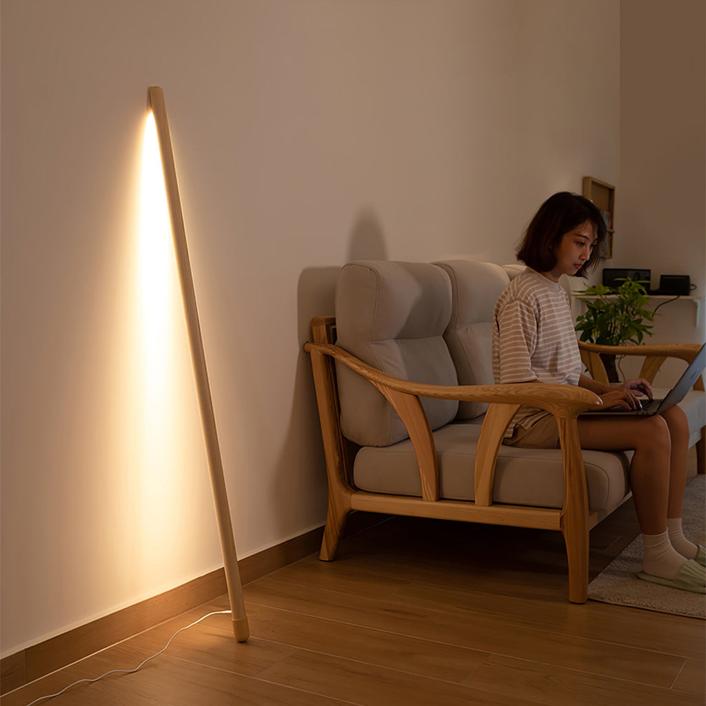 Light wood Clérachic floor lamp leaning in a room corner and lit warm, with a woman on a wooden sofa using a laptop.