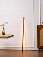 Lit walnut-colour Clérachic floor lamp leaning on a panelled wall, next to a rattan cabinet and a vase of autumn leaves.