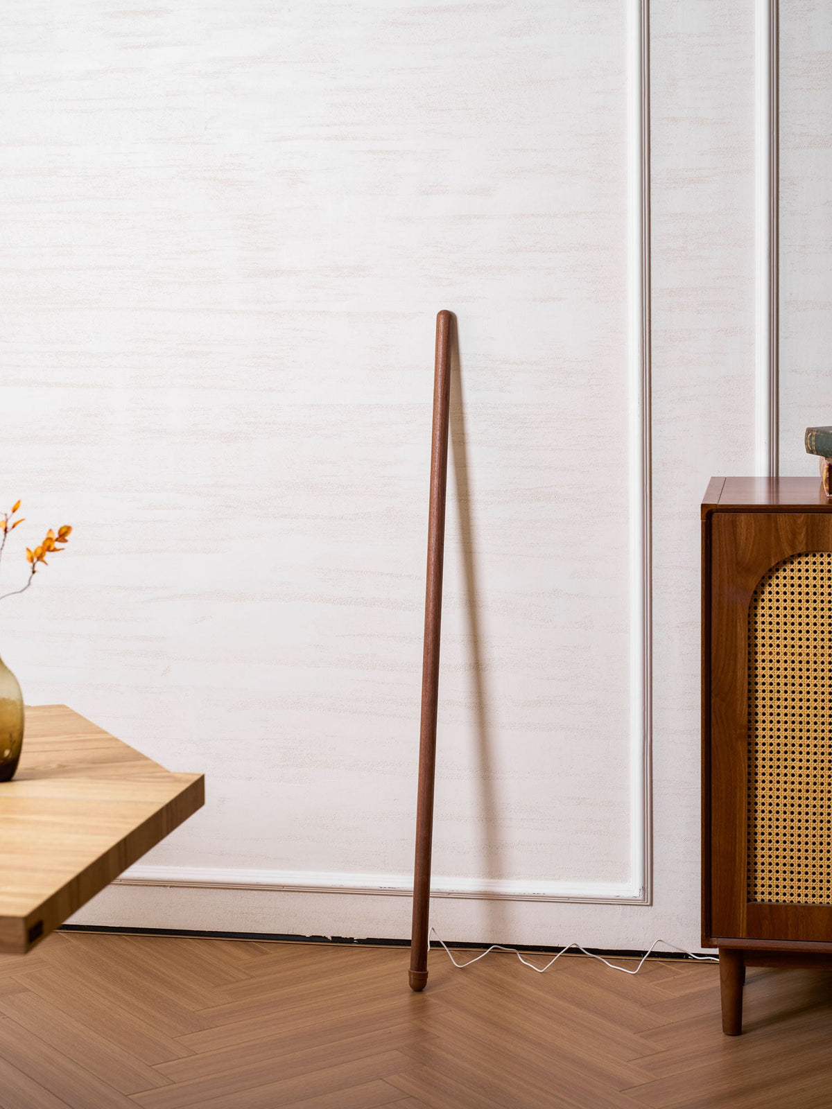 Unlit walnut-colour Clérachic floor lamp leaning on a white panelled wall, beside a pale wood table and a rattan cabinet.