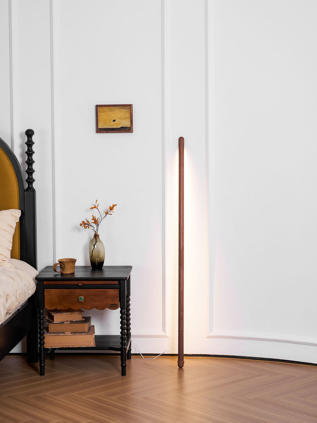 Lit walnut-colour Clérachic floor lamp standing beside a bed and a black bedside table with books and a small vase.