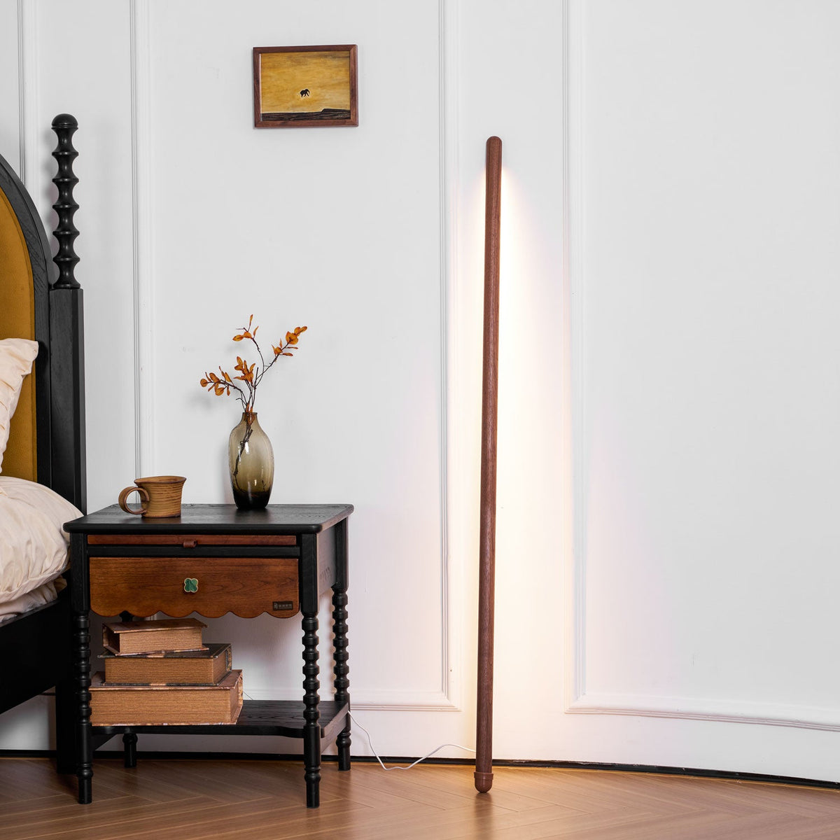 Warm light from a walnut-colour Clérachic floor lamp on a white panelled wall, next to a spindle-legged bedside table.