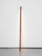 Slim walnut-colour Clérachic floor lamp upright against a pale wall, its light strip casting a soft line of light.