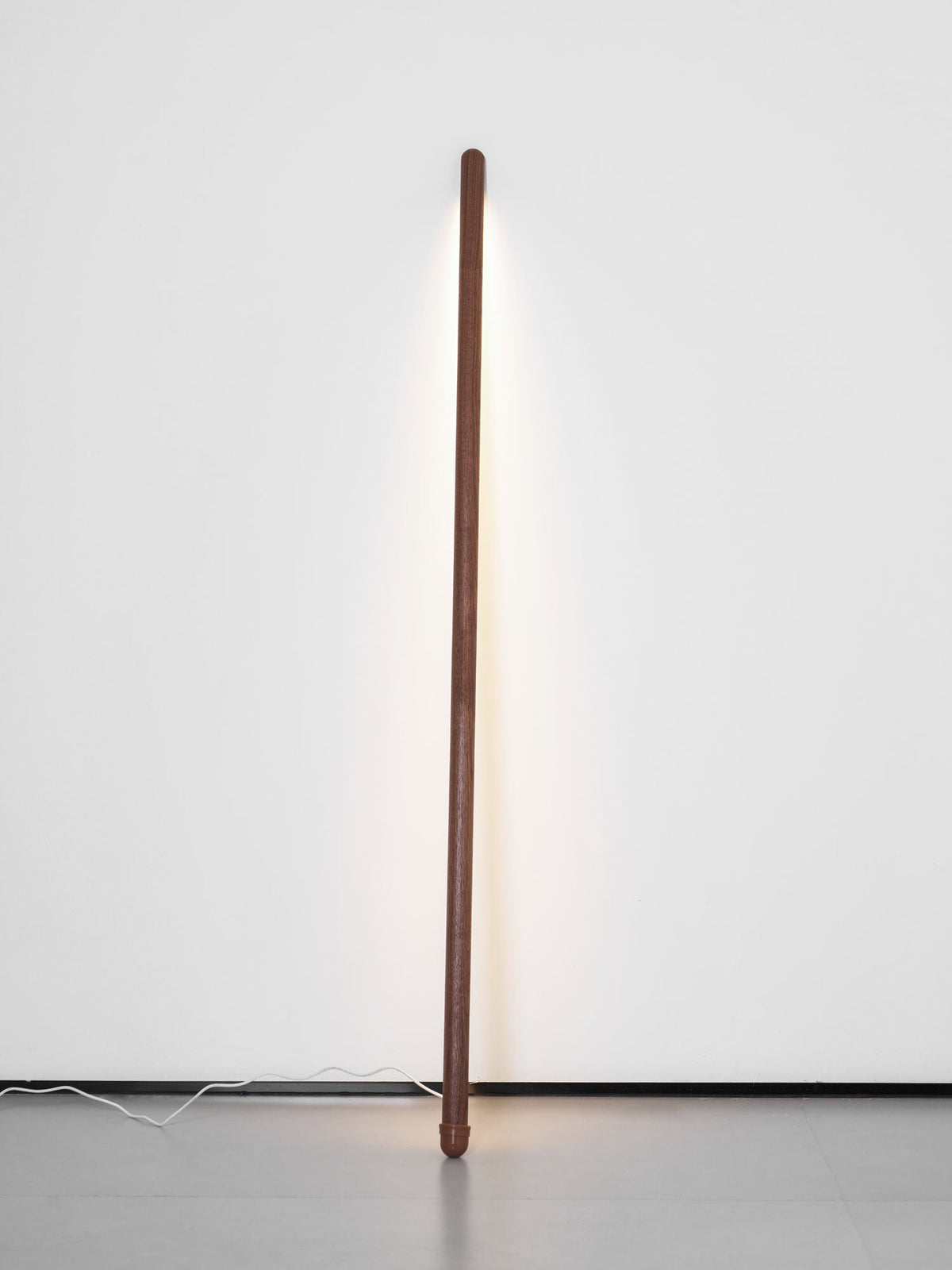 Slim walnut-colour Clérachic floor lamp upright against a pale wall, its light strip casting a soft line of light.