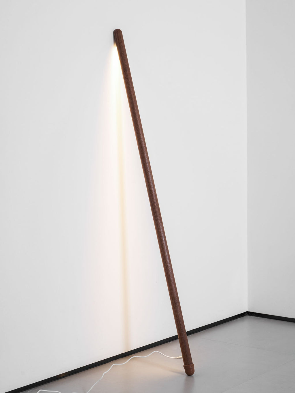 Walnut-colour Clérachic floor lamp leaning in a white corner, lit warm, with a white cord running across the grey floor.