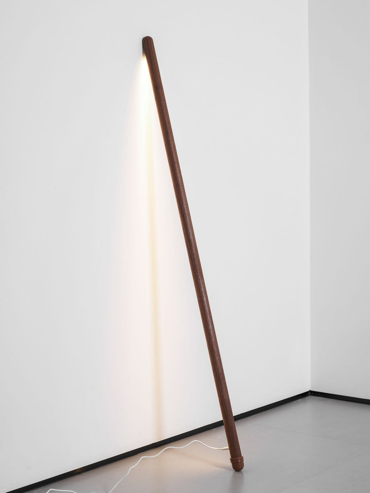 Walnut-colour Clérachic floor lamp leaning in a white corner, lit warm, with a white cord running across the grey floor.