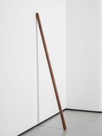 Unlit walnut-colour Clérachic floor lamp leaning in a white corner, with its cord curving across a grey floor.