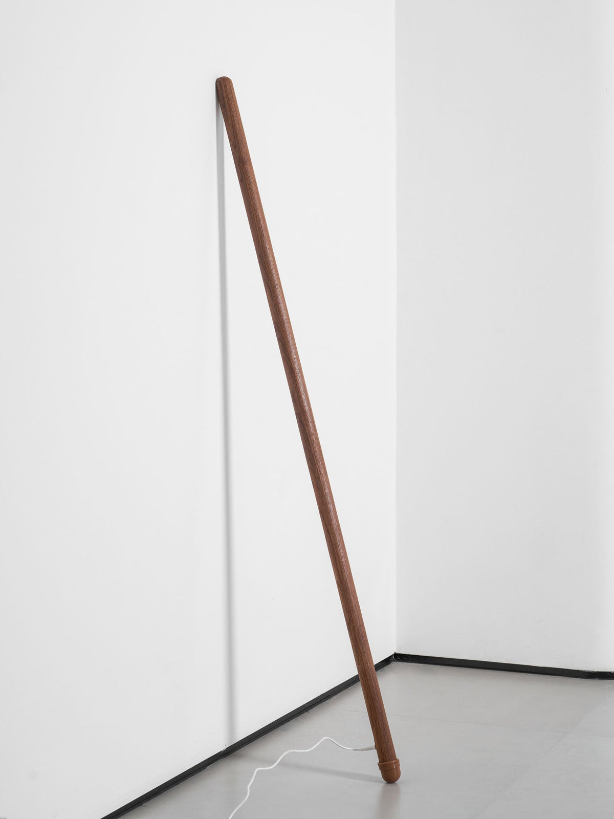 Unlit walnut-colour Clérachic floor lamp leaning in a white corner, with its cord curving across a grey floor.