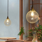 Two Clérachic crackle-glass globe pendants in different sizes hung on twisted cords beside a window and plants.
