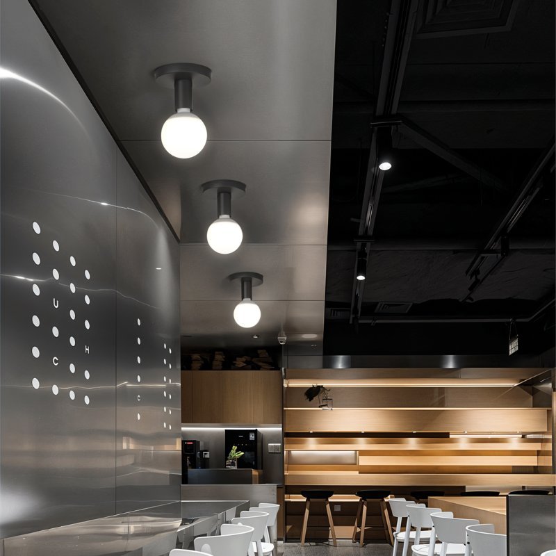 Three Clérachic black ceiling lights with white globes in a row above a steel-clad cafe counter and pale wood seating.