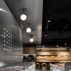Three Clérachic black ceiling lights with white globes in a row above a steel-clad cafe counter and pale wood seating.