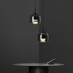 Against a charcoal wall, the Clérachic smoke gray glass pendant hangs in a pair above a dark round table, warm light caught in the glass.