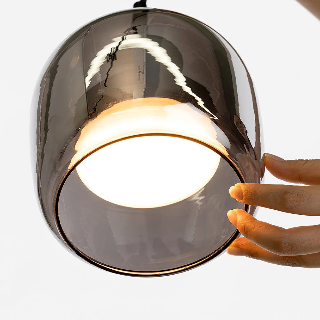 A hand steadies the lit Clérachic pendant shade, the frosted disc bright inside the smoke gray glass and the scale clear against the palm.