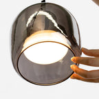 A hand steadies the lit Clérachic pendant shade, the frosted disc bright inside the smoke gray glass and the scale clear against the palm.