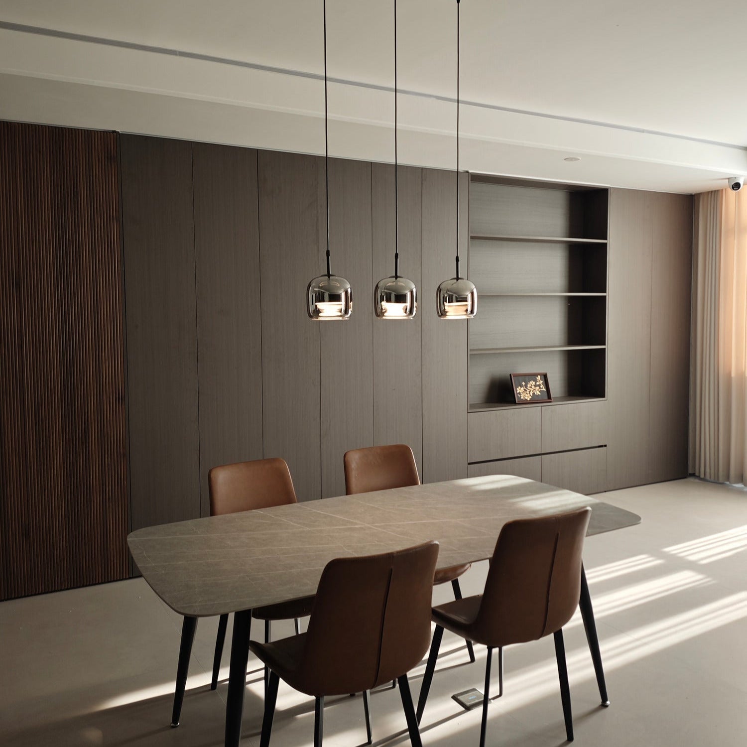 Clérachic smoke gray pendants in a row of three over a stone dining table, lit in a sunlit room with tan leather chairs.