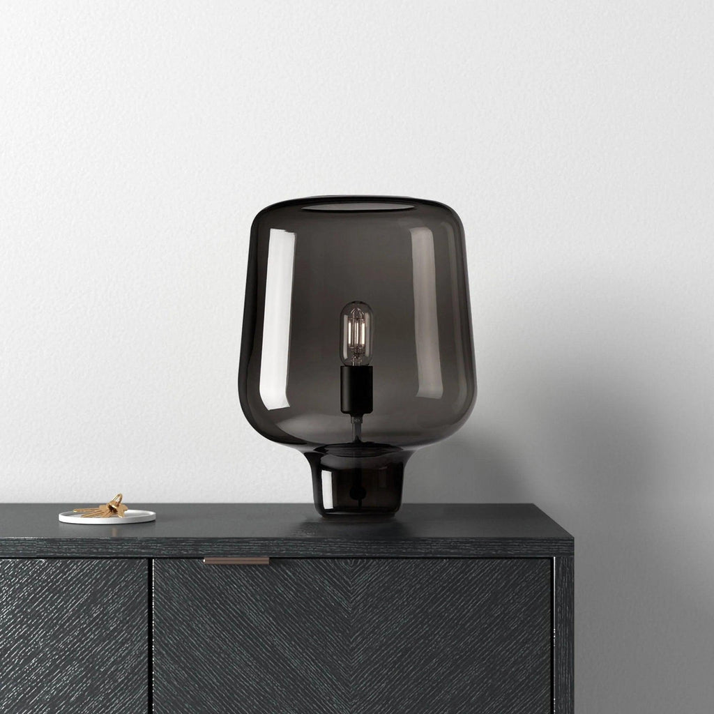 A Clérachic table lamp in smoke grey glass on a black herringbone-front cabinet, against a pale grey wall.