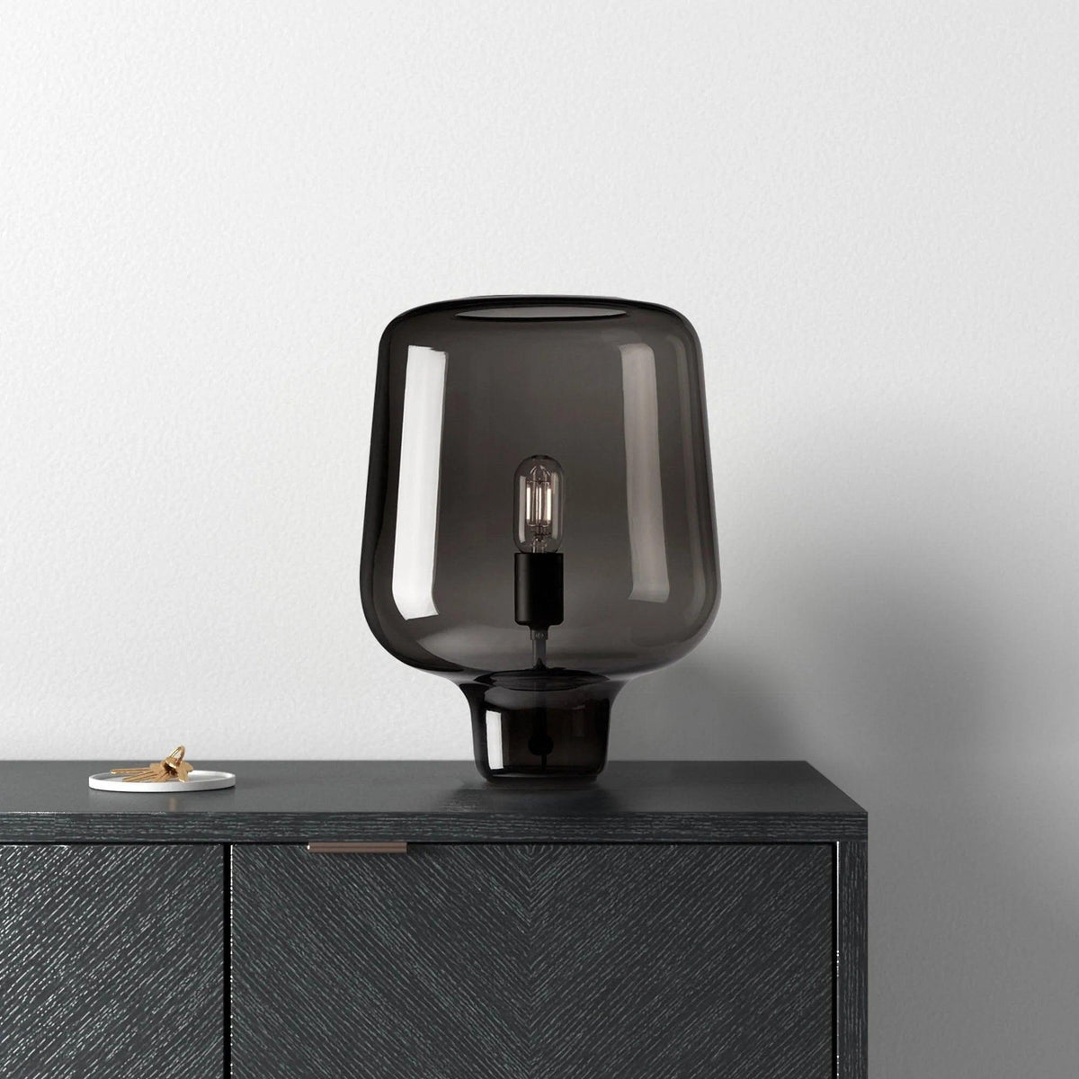 A Clérachic table lamp in smoke grey glass on a black herringbone-front cabinet, against a pale grey wall.