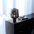 Beside sheer curtains, a Clérachic smoke grey glass lamp glows softly on a black chest of drawers next to two stacked books.