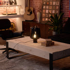 In a dim loft room with brick walls, a lit Clérachic glass table lamp stands on a pale wood coffee table by an armchair.