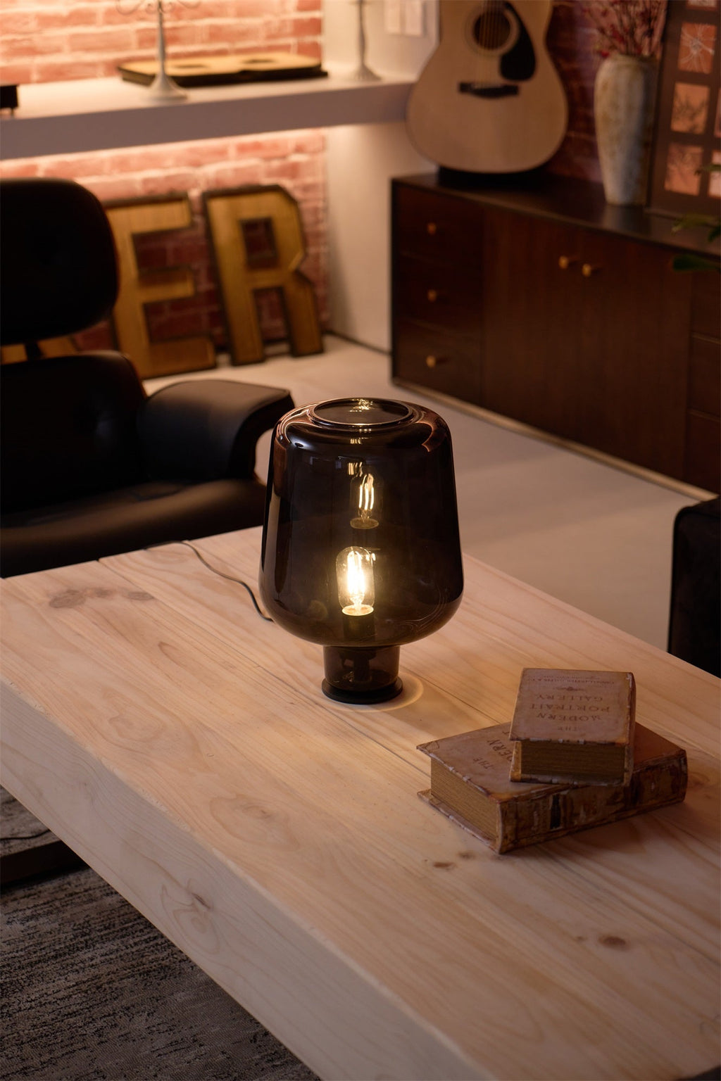 A Clérachic glass table lamp lit on a thick pale wood table, with a stack of old books beside it and a guitar on the wall.
