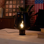 Close view of a lit Clérachic glass table lamp, its smoke grey shade and glowing filament bulb against a brick-wall room.