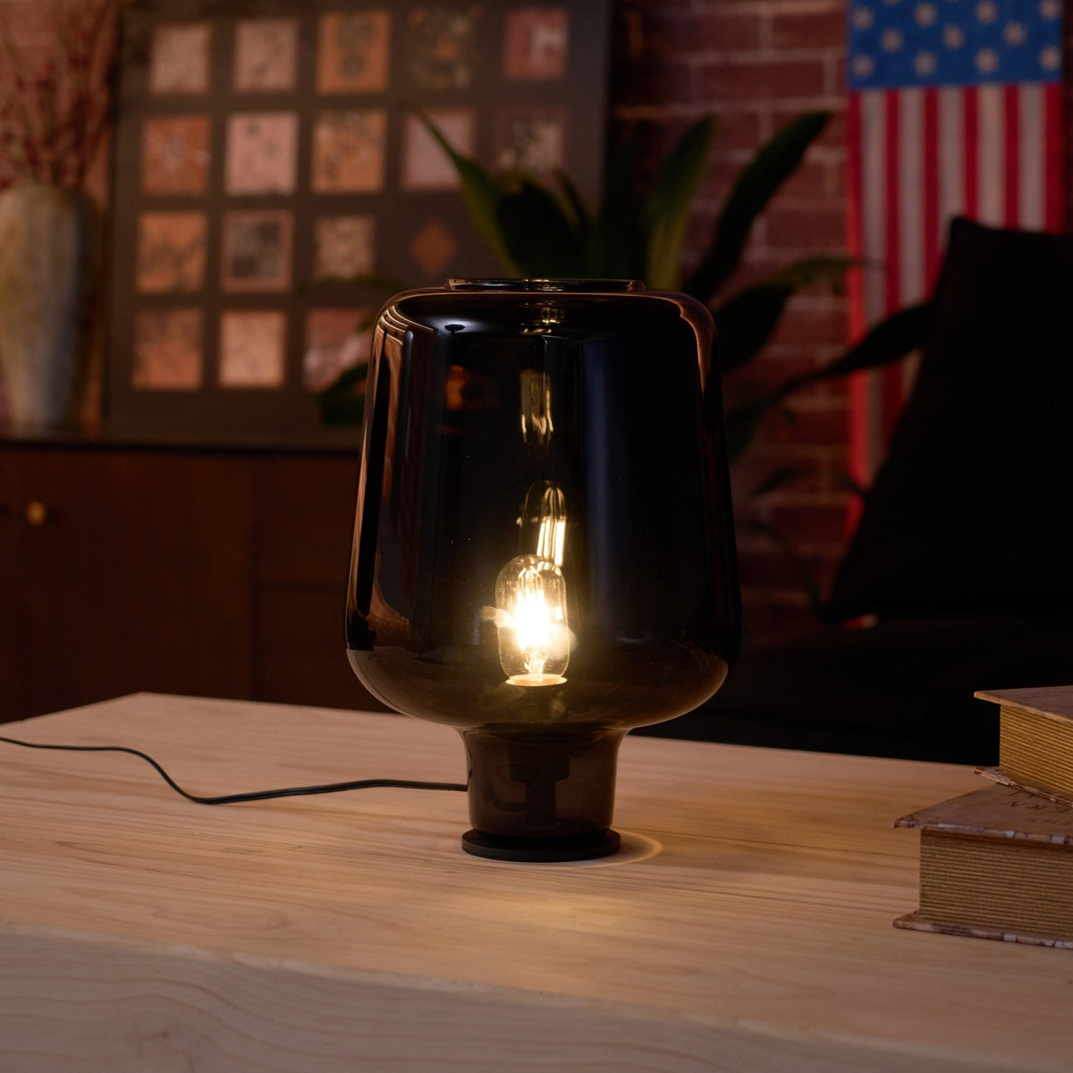 Close view of a lit Clérachic glass table lamp, its smoke grey shade and glowing filament bulb against a brick-wall room.