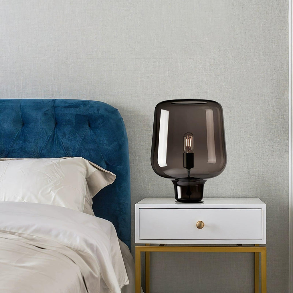 This Clérachic smoke grey glass lamp stands on a white bedside table with a gold base, next to a blue velvet headboard.