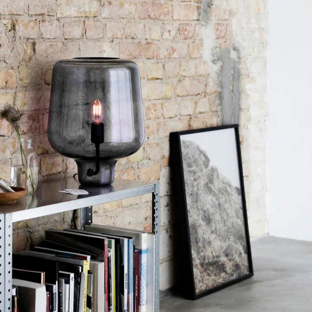 On a metal shelving unit in front of exposed brick, a Clérachic glass table lamp glows pink beside a leaning framed print.